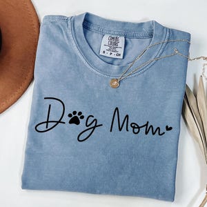 Może przedstawiać: Jasnoniebieska koszulka z napisem "Dog Mom" czarnym pismem, z odciskiem łapy i sercem. Złoty naszyjnik z wisiorkiem jest zarzucony na koszulkę. Koszulka jest złożona.