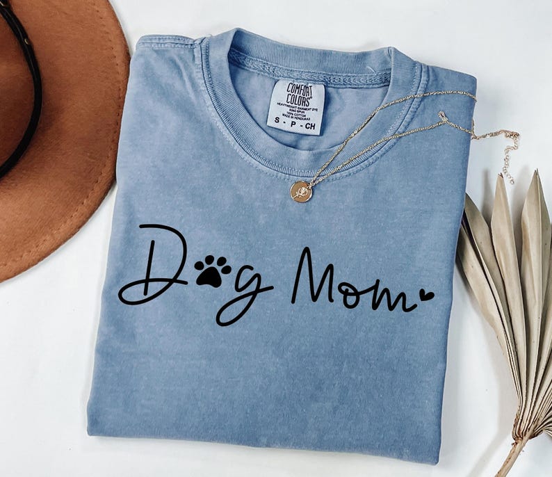 Może przedstawiać: Jasnoniebieska koszulka z napisem "Dog Mom" czarnym pismem, z odciskiem łapy i sercem. Złoty naszyjnik z wisiorkiem jest zarzucony na koszulkę. Koszulka jest złożona.