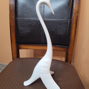 May include: A white ceramic figurine of a long-necked bird, likely a swan or crane, featuring a smooth matte finish, a slender curved neck, and stylized wing details on the sides. The bird stands on a base formed by its own tail and wing tips.