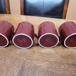 May include: Four maroon ceramic mugs arranged in a row, showing their circular bases. Each base features a white rim and an embossed logo consisting of three interlocking circles above the word ENGLAND, indicating their origin and brand.