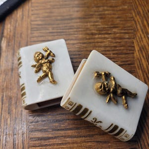 May include: Two small, rectangular, white ceramic boxes with gold accents and a gold cherub design. The boxes are open, revealing the interior. The cherubs are holding arrows and bows. The boxes are on a wooden surface.