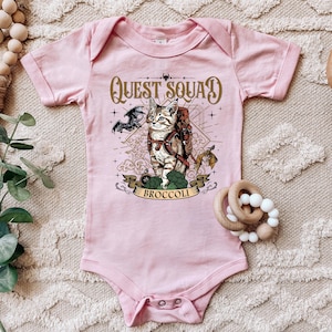 Pode incluir: Macacão rosa para bebê com um gráfico de um gato usando uma mochila e um dragão. O texto "Quest Squad Broccoli" está impresso no macacão.
