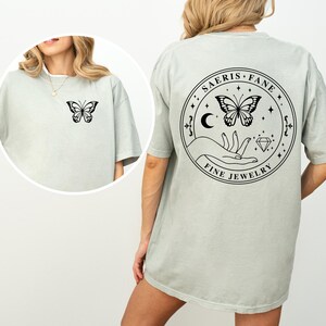May include: A light green t-shirt with a butterfly graphic on the front and back. The back of the shirt features a circular design with a butterfly, hand, moon, and the text "SAERIS FANE FINE JEWELRY".