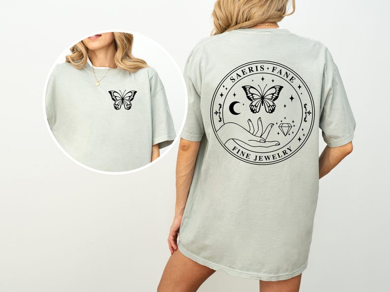 May include: A light green t-shirt with a butterfly graphic on the front and back. The back of the shirt features a circular design with a butterfly, hand, moon, and the text "SAERIS FANE FINE JEWELRY".