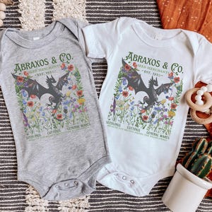 Puede incluir: Dos bodies de bebé con fondo gris y blanco. Ambos bodies presentan un dragón negro rodeado de flores coloridas y el texto "Abraxos & Co. Seed Merchants - Est. 1910 - Seedlings Cuttings General Wares".