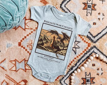 Puede incluir: Un body de bebé gris con un estampado gráfico de un dragón volando sobre un castillo. El texto "Basgiath War College" y "Navarre Morraine Province" está impreso en el body.