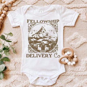 May include: White baby bodysuit with short sleeves. The front features a brown graphic with the text "FELLOWSHIP DELIVERY Co" and a mountain scene. The bodysuit has snap closures at the bottom and is surrounded by wooden toys.