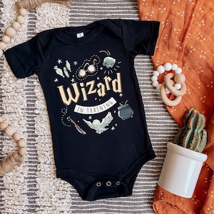Puede incluir: Mono negro para bebé con un búho blanco, gafas y un caldero. El texto en el mono dice "Wizard in Training".
