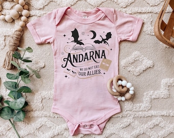 Puede incluir: Body de bebé rosa con la palabra "Andarna" y la frase "We do not eat our allies." El diseño incluye murciélagos, una luna creciente y un libro abierto. Un juguete de madera y cuentas decorativas están en el fondo.