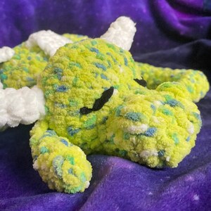 May include: A handmade, plush dragon toy in shades of yellow, green, and blue. The dragon has white horns and wings, and black eyes. The soft, textured material gives the dragon a cuddly appearance.
