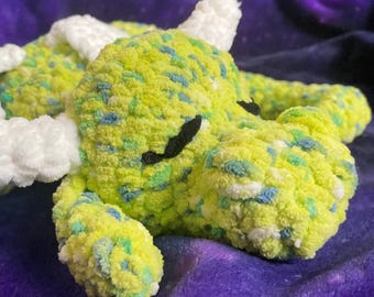 May include: A handmade, plush dragon toy in shades of yellow, green, and blue. The dragon has white horns and wings, and black eyes. The soft, textured material gives the dragon a cuddly appearance.