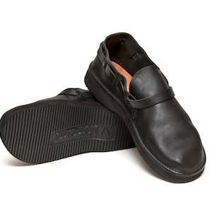 May include: Black leather slip-on shoes with a strap across the top. The soles are thick and textured, with the brand name "Vibram" visible on the sole of one shoe. The shoes are designed for comfort and durability.