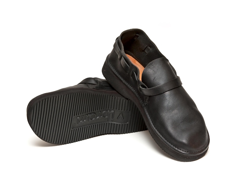 May include: Black leather slip-on shoes with a strap across the top. The soles are thick and textured, with the brand name "Vibram" visible on the sole of one shoe. The shoes are designed for comfort and durability.