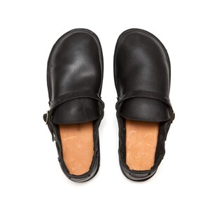 May include: A pair of black leather clogs, viewed from above, with a strap and buckle detail. The interior features a light brown, textured lining. The clogs are set against a white background.