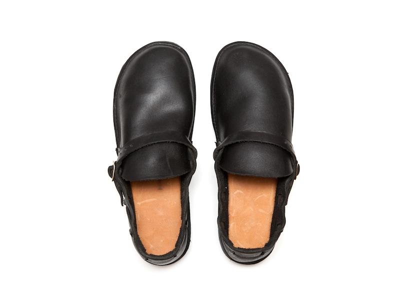 May include: A pair of black leather clogs, viewed from above, with a strap and buckle detail. The interior features a light brown, textured lining. The clogs are set against a white background.