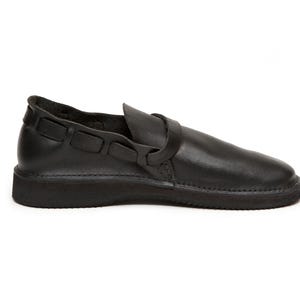 May include: Black leather shoe with a rounded toe and a strap across the top. The shoe has a thick sole and a decorative strap detail on the side. The shoe is a slip-on style.