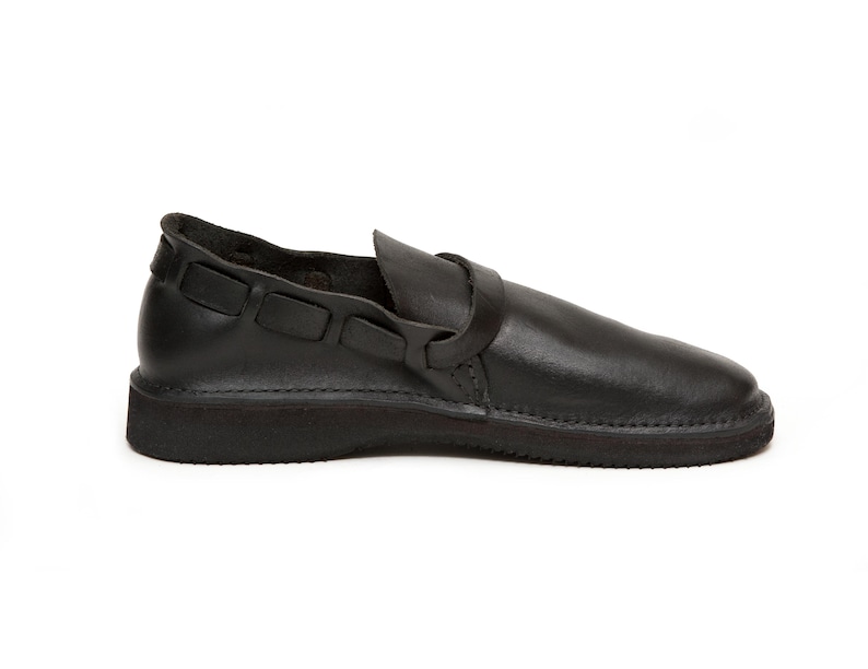 May include: Black leather shoe with a rounded toe and a strap across the top. The shoe has a thick sole and a decorative strap detail on the side. The shoe is a slip-on style.