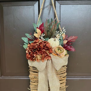 May include: A fall-themed hanging basket with a variety of faux flowers and foliage. The basket is made of woven twigs and features a burlap bow. The arrangement includes orange, red, and green leaves, and a small white pumpkin.