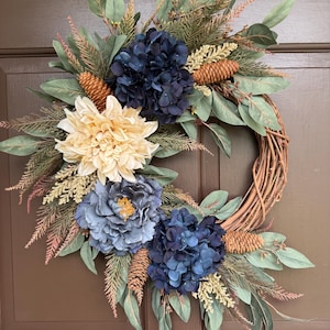 May include: A circular wreath constructed from a woven brown twig base, decorated with dark navy blue hydrangea blossoms, a large cream-colored dahlia, a dusty blue peony, green eucalyptus leaves, feathery foliage, and several small brown pinecones.