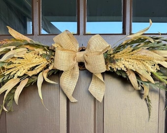 May include: A horizontal door swag featuring a central tan burlap bow surrounded by an arrangement of dried grasses, wheat stalks, and long, slender green and golden-brown leaves. The decorative piece is displayed against a dark brown door.