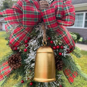 May include: A decorative holiday arrangement featuring a large gold bell suspended by a jute rope. The bell is accented with a red and green plaid ribbon bow, frosted evergreen branches, brown pinecones, and small red berries on a wire frame.