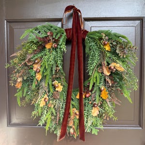 May include: A heart-shaped wreath constructed from green evergreen foliage, featuring scattered brown acorns, small pinecones, and dried yellow-brown leaves. A long, dark burgundy velvet ribbon is tied at the top, hanging down the center.