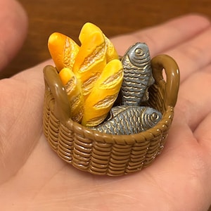 May include: A miniature brown basket containing three golden-yellow loaves of bread and two silver-grey fish. The basket has a woven texture and a handle. The items are set against a blurred background.