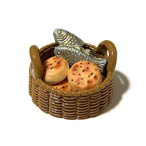 May include: A miniature brown basket containing baked goods and two fish figurines. The basket has a woven design and a handle on each side. The baked goods are round with dark spots, and the fish are silver with gold detailing.