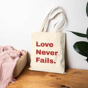 May include: A cream-coloured canvas tote bag featuring the red text Love Never Fails. The bag has two long shoulder straps and a rectangular body. The design is printed in a bold, sans-serif font centred on the front of the fabric tote bag.