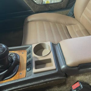 May include: Interior car detail featuring a tan leather seat and center console. The console includes a cup holder, storage compartment, and wooden trim. A black gear shift and seatbelt buckle are also visible.
