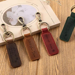 May include: Leather keyrings in brown, tan, red, and green with metal fixings. Each keyring has a rectangular tag with engraved text and designs. The text includes dates, phrases, and a sun symbol. The keyrings are on a wooden surface.