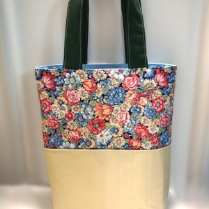 May include: A floral tote bag with a cream-colored bottom and dark green handles. The upper portion of the bag features a colorful floral pattern with red, blue, and yellow flowers. The handles are a solid dark green.