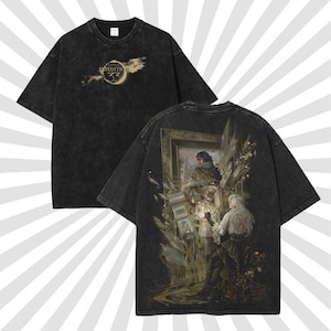 May include: Black t-shirt with a vintage wash effect. The front features the text "EXPEDITION 33" with a crescent moon design. The back of the shirt displays a detailed illustration of figures in a framed artwork, with a soft, faded color palette.