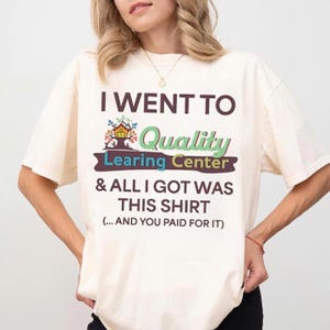 May include: Cream-colored t-shirt with the text "I WENT TO Quality Learing Center & ALL I GOT WAS THIS SHIRT (...AND YOU PAID FOR IT)". The shirt has a crew neck and short sleeves. The text is in various colors and fonts.