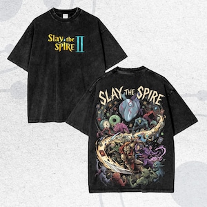 May include: Black t-shirt with the text "Slay the Spire II" on the front. The back of the shirt features a colorful graphic of characters and creatures from the game "Slay the Spire".