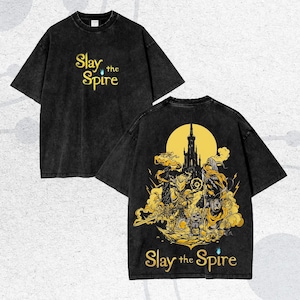 May include: Black t-shirt with the text "Slay the Spire" in gold. The back of the shirt features a detailed gold illustration of a castle, characters, and the same text. The shirt has a vintage, washed look.