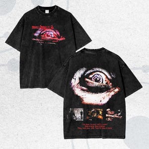 Peut inclure: Deux t-shirts noirs surdimensionnés avec un motif graphique vieilli. Le devant du t-shirt présente le texte "manhunt 2" au-dessus d'un œil stylisé. Le dos du t-shirt affiche un gros plan d'un œil et le texte "The door to your cell is open. One choice. One chance. They took your life. Time to take it back."