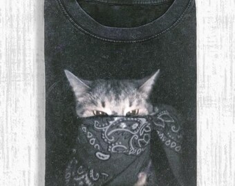 May include: Dark gray t-shirt featuring a cat wearing a black bandana with a paisley design. The cat's eyes are visible above the bandana. The shirt is folded and has a crew neck.