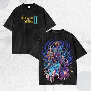 May include: Black t-shirt with the text "Slay the Spire II" in yellow and teal. The back of the shirt features a colorful graphic of fantasy characters in shades of blue, purple, and yellow.