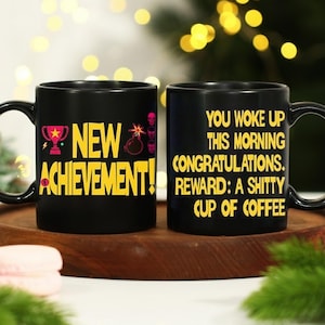 以下が含まれることがあります： ユーモラスなテキストが入った2つの黒いセラミックマグカップ。1つのマグカップには、トロフィーと爆弾のグラフィックとともに、黄色で「NEW ACHIEVEMENT！」と書かれています。もう1つは「YOU WOKE UP THIS MORNING. CONGRATULATIONS. REWARD: A SHITTY CUP OF COFFEE.」と書かれています。
