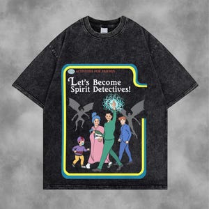 Peut inclure: T-shirt gris foncé avec un motif graphique représentant le texte "Let's Become Spirit Detectives !" et des illustrations de personnes et de figures ailées. Le motif est encadré d'une bordure jaune et bleue, avec les mots "ACTIVITÉS POUR AMIS" au-dessus du texte principal.