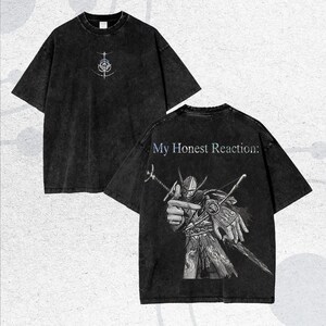 May include: Two-sided, dark gray t-shirts. The front features a white and light blue graphic. The back displays a detailed illustration of a warrior with the text "My Honest Reaction:" in light blue.