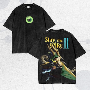 May include: Black t-shirt with a distressed wash. The front features a green and yellow circular graphic. The back displays the text "Slay the Spire II" above a detailed illustration of a character with horns and a green cloak.
