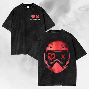 Könnte beinhalten: Schwarzes T-Shirt im Used-Look mit roten Grafiken. Auf der Vorderseite ein Herz und ein X mit dem Text "CAUGHT UP". Auf der Rückseite ein rotes Helmdesign mit Herz und X. Streetwear-Stil.