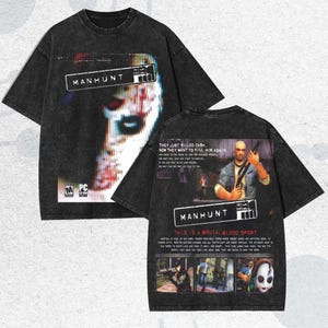 Puede incluir: Camiseta gris oscuro con gráficos del videojuego "Manhunt". La parte delantera presenta una cara distorsionada y el logotipo del juego. La parte trasera muestra un personaje con un arma y otras imágenes del juego. La camiseta tiene un aspecto desgastado.