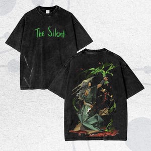 May include: Black oversized t-shirt with the text "The Silent" in green. The back of the shirt features a detailed illustration of a figure in green robes holding a green plant, with red and green accents. The shirt has a vintage, washed look.