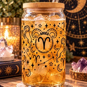 May include: A clear glass tumbler with a bamboo lid and glass straw, filled with an amber liquid and ice. The exterior features black printed graphics of the Aries zodiac symbol, a ram, stars, and moons, with the text ARIES, CONFIDENT, COURAGEOUS, ADVENTUROUS.