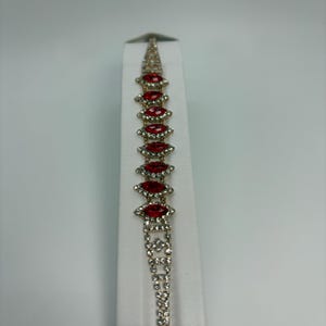 May include: A metallic bracelet featuring a series of marquise-cut red stones, each surrounded by a border of small clear rhinestones. The bracelet is constructed with a flexible link design, incorporating additional clear rhinestone accents.
