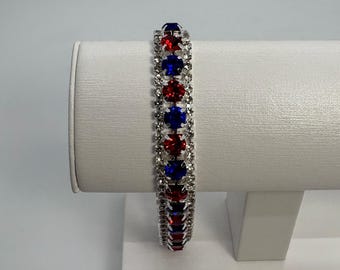 May include: A silver-toned bracelet featuring a central row of alternating round red and blue faceted stones, flanked by two outer rows of small clear crystals. The design is symmetrical, with the stones set in a metallic frame that catches light.