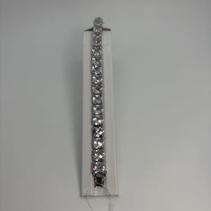 May include: A silver-toned bracelet with a series of square-cut, clear gemstones. The bracelet is displayed on a white stand against a plain background. The gemstones are evenly spaced and reflect light, creating a sparkling effect. The clasp is visible at the bottom.